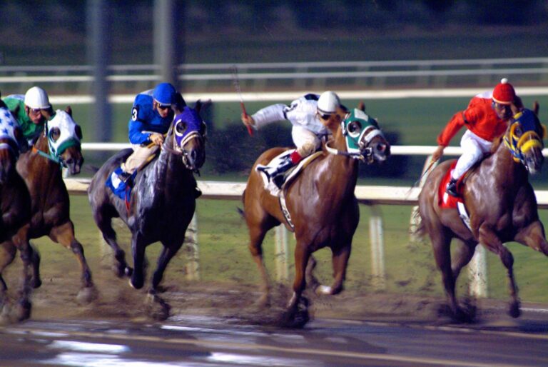 Three jockeys racing horses on a dirt track under lights.